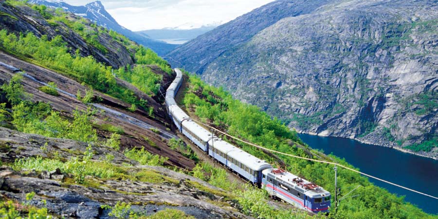 A long, light blue train travels along a cliffside. 